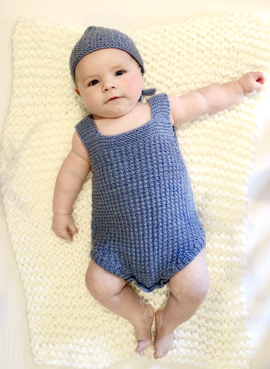 A baby in a knitted blue romper and bonnet lying on a cream knitted blanket