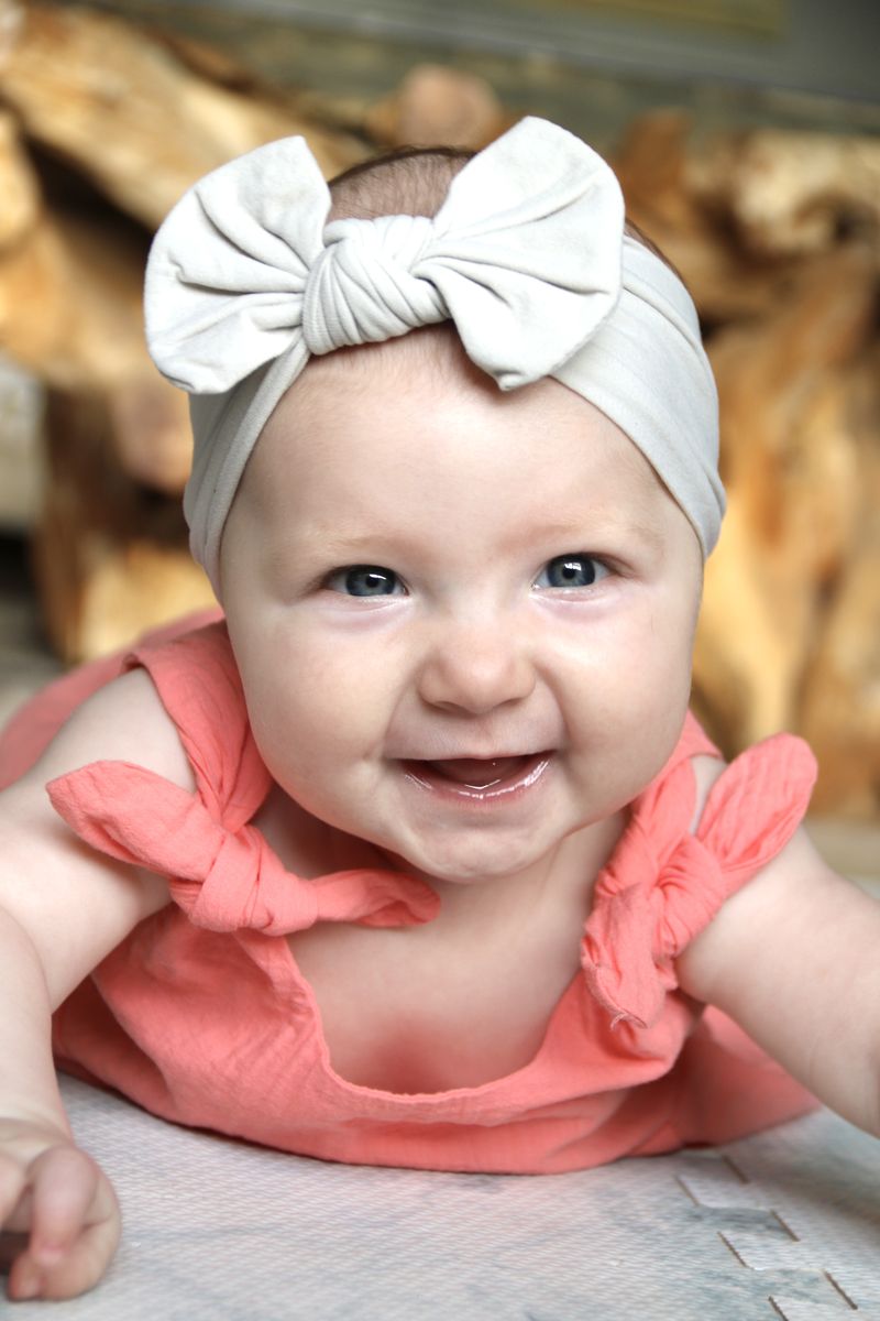 A laughing baby in a coral top and grey bow headband during tummy time