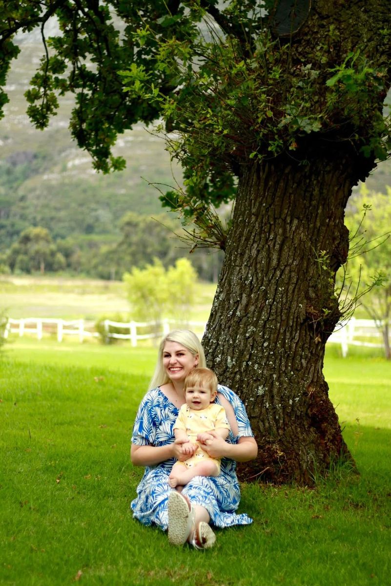 A smiling mother sitting on the grass under a large tree with her baby on her lap