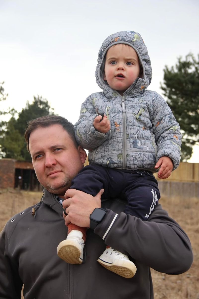 A toddler in a dinosaur-print puffer jacket riding on his dad's shoulders