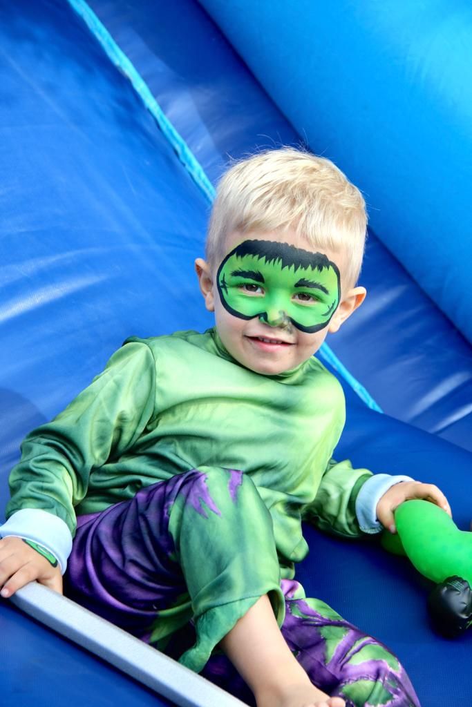A boy in a green superhero costume and face paint on a blue jumping castle slide
