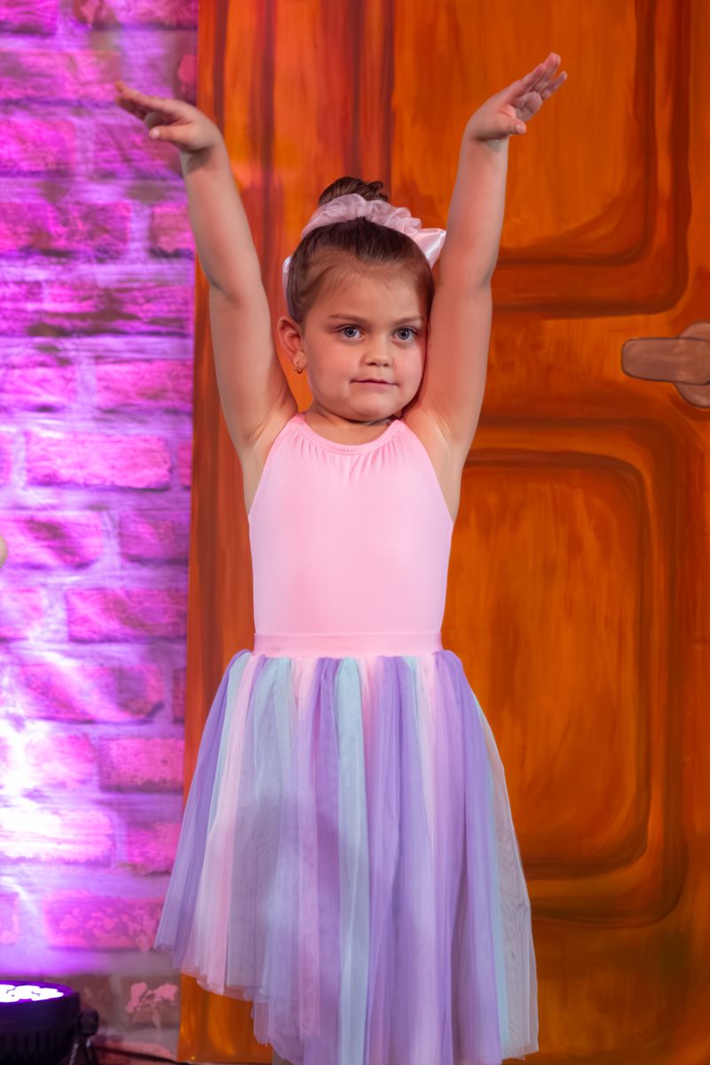 A girl in a pink leotard and pastel tutu with both arms raised on stage